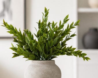 Könnte beinhalten: Ein Strauß aus künstlichen grünen Blattstielen, arrangiert in einer strukturierten, hellgrauen Keramikvase. Die Pflanze besteht aus zahlreichen kleinen, spitzen Blättern an mehreren verzweigten Stielen, die aus der Mitte des Gefäßes ragen.