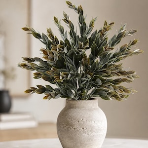 May include: A beige, textured, round ceramic vase with a narrow neck holds a dense arrangement of artificial green and brownish-tipped leafy branches. The plant features numerous small, pointed leaves extending outward in a full, rounded shape.