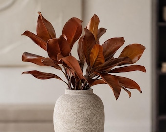 May include: A round, textured, light gray stone vase containing a cluster of deep copper-brown magnolia leaves. The leaves are broad, smooth, and arranged in a dense, upright display, contrasting with the matte, speckled surface of the vase.