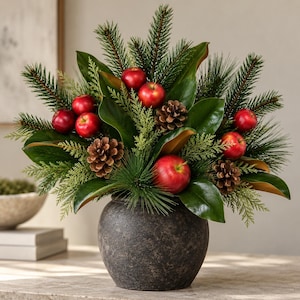 May include: A decorative arrangement featuring artificial red apples, brown pinecones, green magnolia leaves, and evergreen pine needles. The botanical display is contained within a round, dark gray, textured ceramic vase with a matte finish.