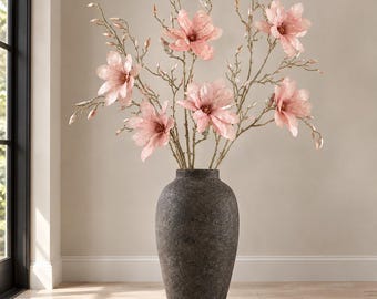 May include: A tall, dark gray, textured floor vase holds a large arrangement of light pink magnolia flowers on slender, branching stems. The vase has a rounded body and a narrow neck, while the flowers feature delicate, layered petals.