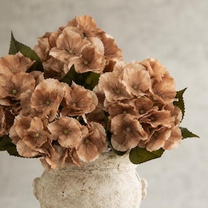 May include: A bouquet of artificial hydrangea flowers in muted shades of dusty brown and tan, featuring layered petals and dark green leaves. The flowers are arranged in a textured, off-white ceramic vase with a rounded body and small handles.