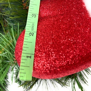 One of the red glittered mittens from the floral spray on a Christmas pine background with a ruler showing width of 4".