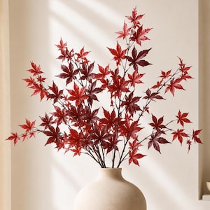 May include: A decorative arrangement of artificial maple branches featuring vibrant red and deep burgundy leaves. The slender, dark brown stems are gathered together and displayed in a smooth, rounded, light beige ceramic vase.