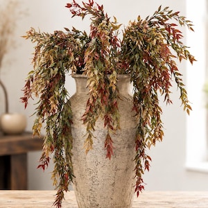 May include: A tall, textured, off-white ceramic vase with a tapered neck and a wide base, filled with cascading artificial foliage. The plant stems feature a mix of autumn-toned leaves in shades of deep red, burnt orange, and muted olive green.