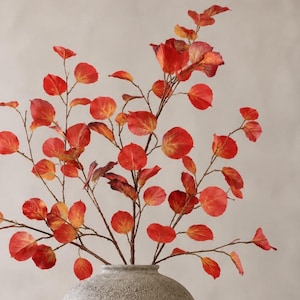 May include: A decorative branch with vibrant red and orange circular leaves arranged on thin brown stems, displayed in a textured, light gray spherical vase. The foliage features autumn-inspired colors with visible veins on each leaf surface.