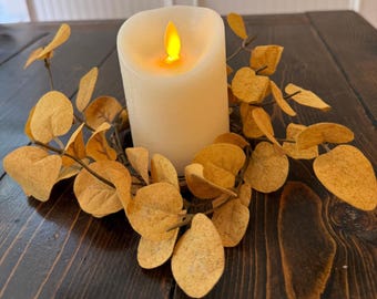 May include: A cream-colored LED candle with a flickering amber flame sits atop a decorative wreath of golden-brown leaves. The candle is placed on a dark brown wooden surface, creating a warm, inviting ambiance. This item is perfect for home decor.