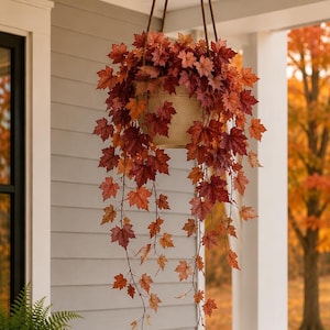May include: A hanging planter containing artificial maple vines with vibrant red, orange, and deep burgundy leaves. The plant is suspended by three brown ropes attached to a neutral-colored, round pot, creating a cascading display of autumn foliage.