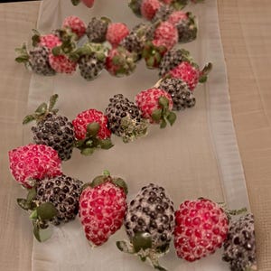 May include: A close-up of a decorative garland featuring artificial strawberries and blackberries. The berries are in varying shades of red and black, with green leaves and a frosted appearance. The garland is arranged in a curved pattern.