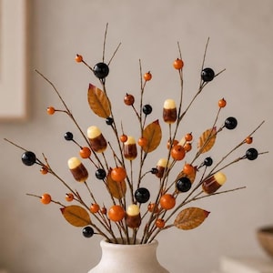 May include: A decorative floral arrangement featuring thin brown branches adorned with glossy black berries, bright orange berries, small brown leaves, and acorn-shaped ornaments with cream-colored tops and brown bases, all set in a light-colored vase.