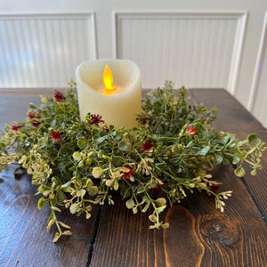 May include: A white pillar candle with a flickering flame sits in the center of a green and red artificial floral wreath.
