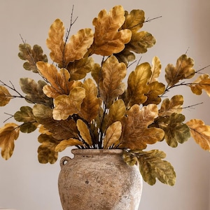 May include: A rustic, textured, light brown ceramic vase with two small side handles holds a large, dense arrangement of artificial oak branches. The branches feature numerous lobed leaves in varying shades of golden yellow, amber, and muted olive green.