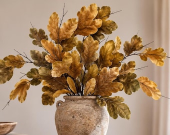 May include: A rustic, textured, light brown ceramic vase with two small side handles holds a large, dense arrangement of artificial oak branches. The branches feature numerous lobed leaves in varying shades of golden yellow, amber, and muted olive green.