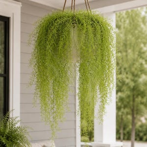 May include: A hanging basket with cascading, bright green artificial foliage. The plant is suspended by brown rope from a porch ceiling. The basket is white, and the background shows a porch with gray siding and outdoor furniture.