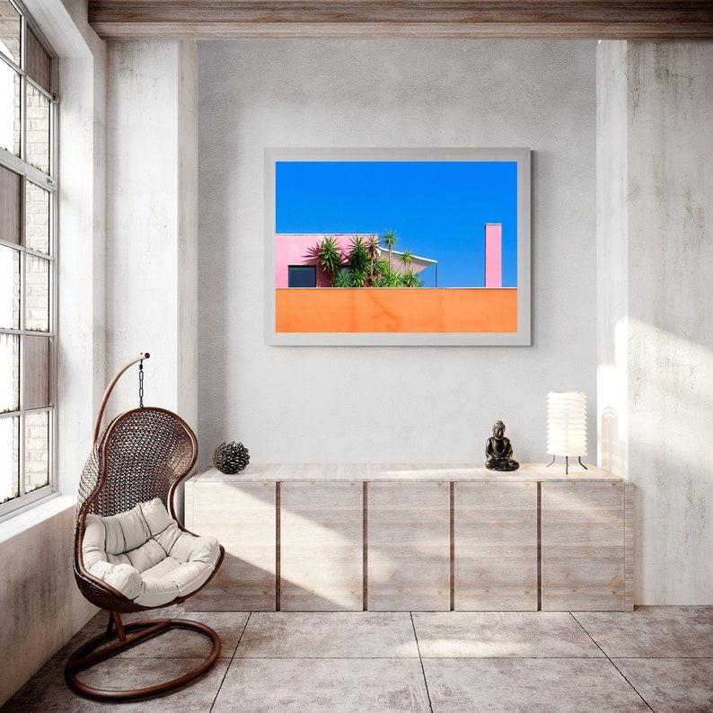 May include: A framed print featuring a vibrant architectural scene. The artwork showcases a building with pink and orange walls, lush green plants, and a bright blue sky. The print is displayed in a modern, minimalist interior with a hanging chair and wooden cabinet.