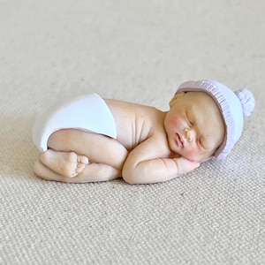 May include: A small, sleeping baby doll figurine with a white diaper and a lavender knitted hat. The doll is lying on a white textured surface.