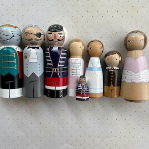 May include: A set of eight wooden peg dolls, painted to resemble characters from the Nutcracker ballet. The dolls are dressed in colorful costumes and have painted facial features. The dolls are arranged in a row on a white background.