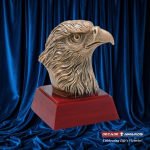 May include: A bronze eagle head sculpture on a red wooden base. The eagle has a detailed, textured design and is looking to the right. The base is square and has a smooth finish. The sculpture is on a blue velvet background.