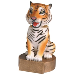 May include: A figurine of a tiger sitting on a bronze-colored base. The tiger has orange and black stripes and a white chest. The tiger is looking to the right and has its mouth open.