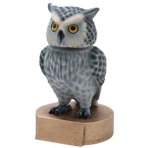 May include: A gray and white owl figurine with yellow eyes, perched on a bronze-colored base. The owl is detailed with feathers and has a realistic appearance.