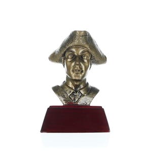 May include: A bronze bust of a person wearing a hat, on a red wooden base. The bust is of a person with a serious expression, and the hat is a tri-cornered hat.