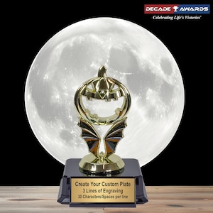 May include: A gold-colored Halloween-themed trophy with a pumpkin-shaped top and bat wing accents, set against a full moon backdrop. The trophy sits on a black base with a plate that reads "Create Your Custom Plate, 3 Lines of Engraving, 30 Characters/Spaces per line."