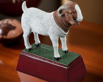 May include: A white and brown goat figurine with horns standing on a maroon and green base. The figurine is a trophy or award.