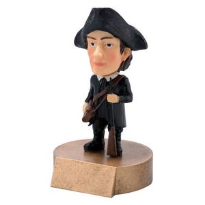 May include: A small figurine of a man wearing a black tricorn hat and a black coat, holding a rifle. He is standing on a gold-colored base.