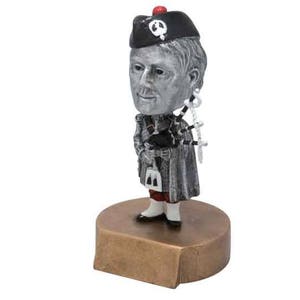 May include: A silver figurine of a person wearing a black hat with a white skull and crossbones design, playing a bagpipe. The figurine is standing on a round bronze base.
