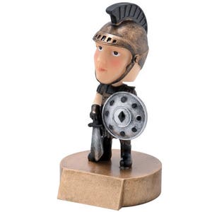 May include: A small figurine of a Roman soldier wearing a bronze helmet and holding a sword and shield. The figurine is standing on a gold-colored base.