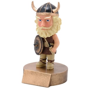 May include: A small figurine of a Viking warrior wearing a horned helmet and holding a shield. The figurine is made of brown and gold painted resin and is standing on a gold base.