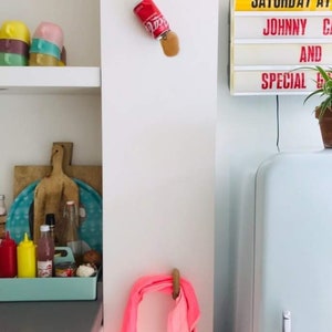 Op de afbeelding: Een witte keuken met een vintage koelkast, een roze handdoek die aan een haak hangt en een bord met de tekst "SATURDAY AT JOHNNY'S AND SPECIAL GUESTS".