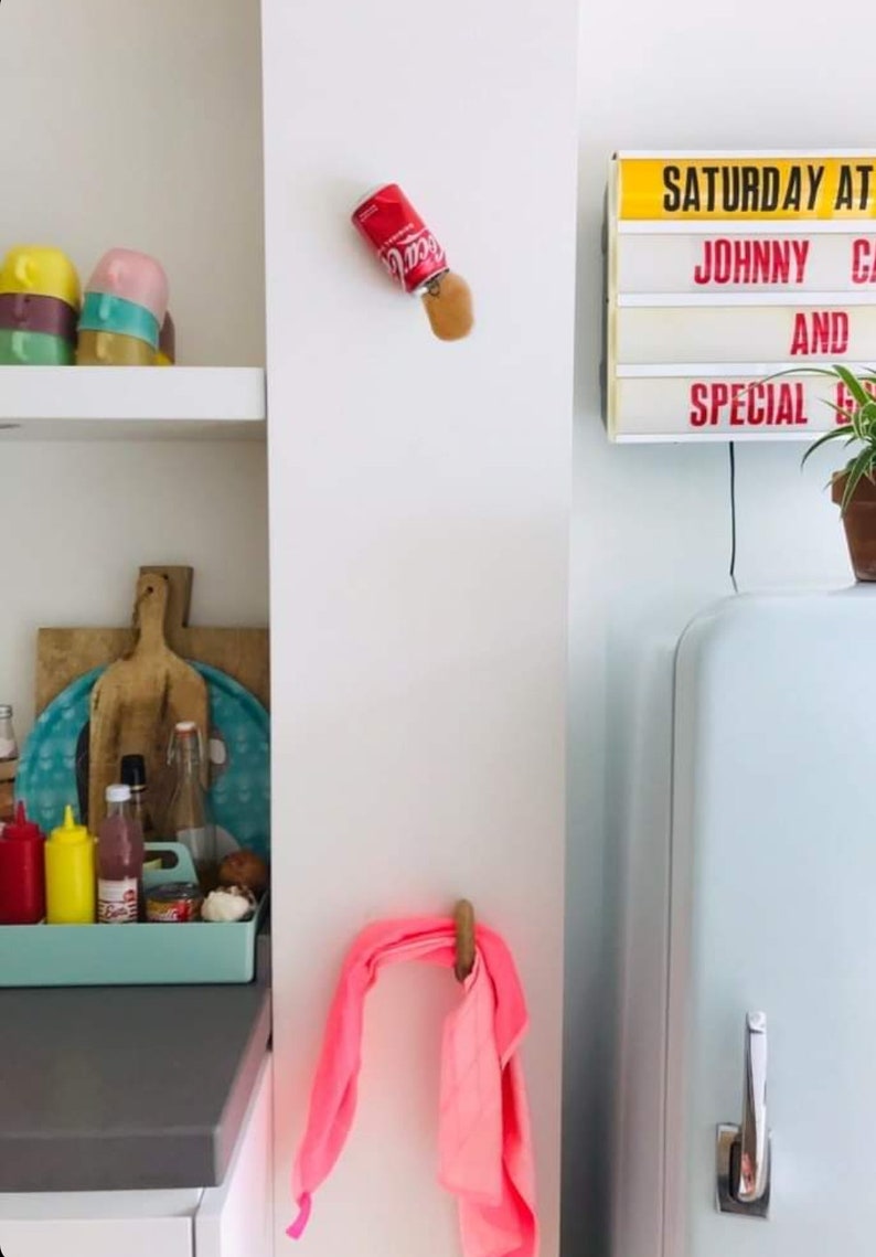 Op de afbeelding: Een witte keuken met een vintage koelkast, een roze handdoek die aan een haak hangt en een bord met de tekst "SATURDAY AT JOHNNY'S AND SPECIAL GUESTS".