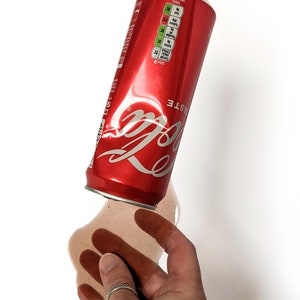 Op de afbeelding: Een rode aluminium blikje Coca-Cola met het logo en de woorden "Taste the Feeling" erop gedrukt. Het blikje ligt op zijn zijkant en morst bruine vloeistof op een wit oppervlak.