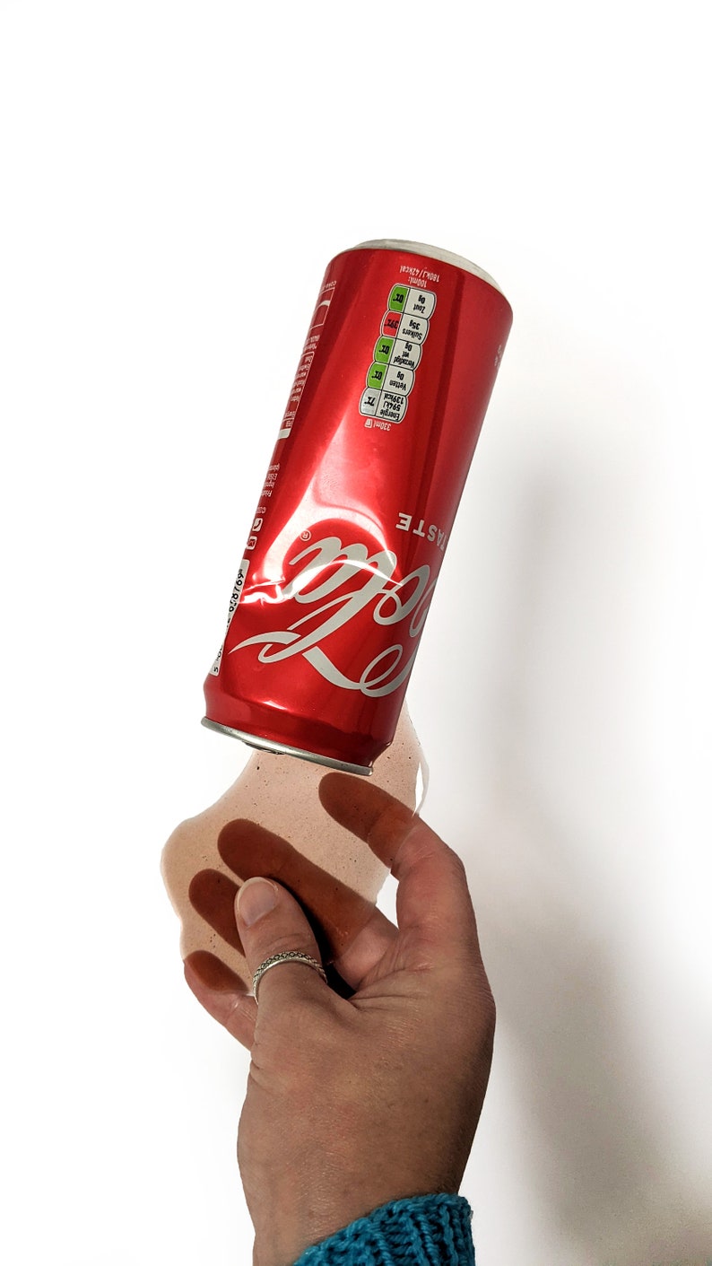 Op de afbeelding: Een rode aluminium blikje Coca-Cola met het logo en de woorden "Taste the Feeling" erop gedrukt. Het blikje ligt op zijn zijkant en morst bruine vloeistof op een wit oppervlak.