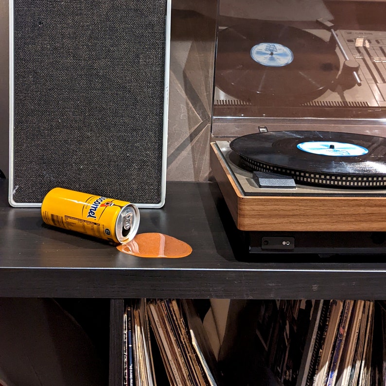 Op de afbeelding: Een gele blikje frisdrank met het woord "Corona" erop gedrukt, gemorst op een zwart oppervlak. Het blikje ligt naast een vintage platenspeler met een houten basis.