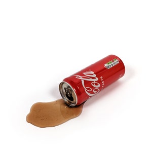 Op de afbeelding: Een rode aluminium blikje Coca-Cola met de woorden "Coca-Cola" en "Taste" erop gedrukt. Het blikje ligt op zijn zijkant met een bruine vloeistof die eruit loopt.