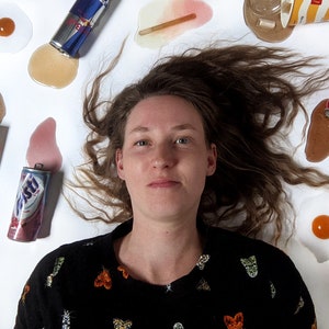 Op de afbeelding: Een jonge vrouw met lang bruin haar ligt op een wit oppervlak omringd door verschillende gemorst drinken en voedsel. De items omvatten koffie, frisdrank, bier en eieren. De vrouw draagt een zwarte trui met een kleurrijk patroon.