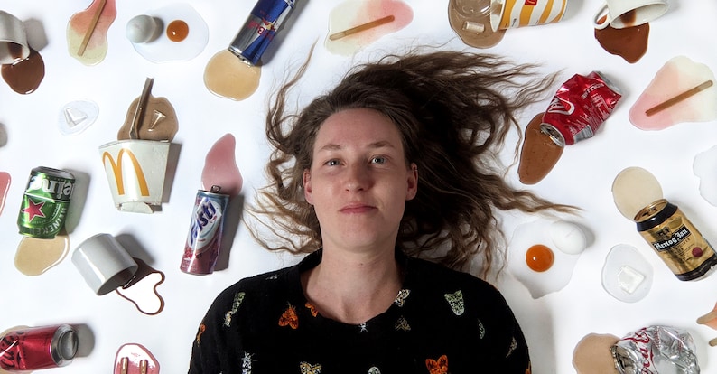 Op de afbeelding: Een jonge vrouw met lang bruin haar ligt op een wit oppervlak omringd door verschillende gemorst drinken en voedsel. De items omvatten koffie, frisdrank, bier en eieren. De vrouw draagt een zwarte trui met een kleurrijk patroon.