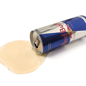 Peut inclure: Une canette argentée de boisson énergisante Red Bull a été renversée, répandant le liquide jaune sur une surface blanche. La canette est couchée sur le côté, avec le logo et le texte Red Bull visibles.