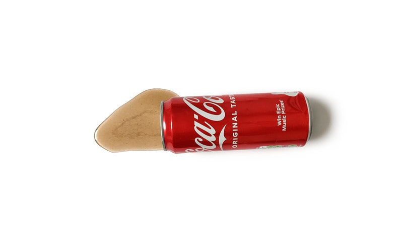 Op de afbeelding: Een rode Coca-Cola blikje met de tekst "ORIGINAL TASTE" en "Win Epic Music Prizes" erop gedrukt. Het blikje ligt op zijn zijkant en de vloeistof loopt eruit.