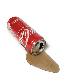 Op de afbeelding: Een rode blik Coca-Cola met de woorden "Coca-Cola" en "Originele Smaak" erop gedrukt ligt op zijn zij op een wit oppervlak. De blik lekt bruine vloeistof.