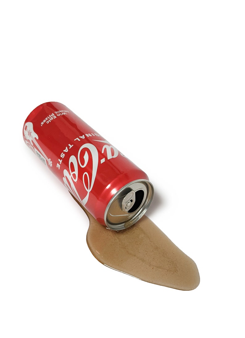 Op de afbeelding: Een rode blik Coca-Cola met de woorden "Coca-Cola" en "Originele Smaak" erop gedrukt ligt op zijn zij op een wit oppervlak. De blik lekt bruine vloeistof.