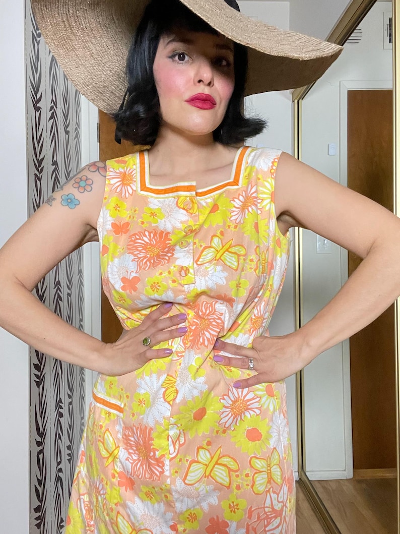 May include: A sleeveless dress with a floral print in shades of yellow, orange, and white. The dress has a square neckline and a button-up front. The model is wearing a wide-brimmed straw hat.