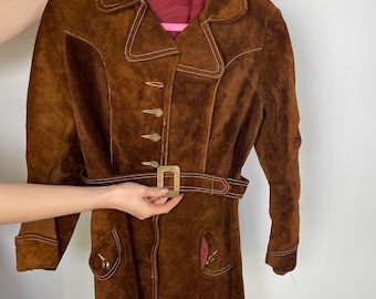 May include: A brown suede jacket with a belt and button closure. The jacket has a collar and long sleeves.