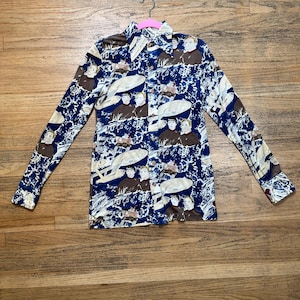 Peut inclure: Une chemise à manches longues bleue avec un imprimé de vaisseau spatial et de multiples personnages. La chemise a un devant boutonné et un col.