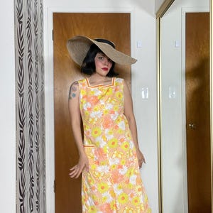 May include: A sleeveless yellow and orange floral dress with a white trim and a large pocket on the left side. The dress is worn with a wide-brimmed straw hat.