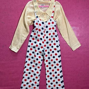 May include: White polka dot jumpsuit with red and blue polka dots. The jumpsuit has thin straps and a scoop neck. A yellow button-up shirt with a collar is layered over the jumpsuit.