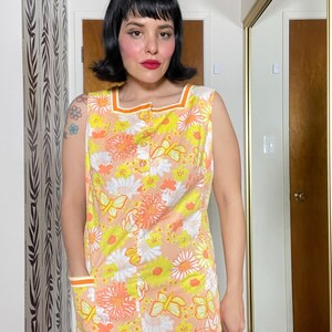 May include: A sleeveless dress with a floral print in shades of yellow, orange, and white. The dress has a button-up front and two pockets on the sides. The dress has a contrasting orange trim around the neckline and armholes.