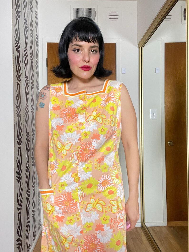 May include: A sleeveless dress with a floral print in shades of yellow, orange, and white. The dress has a button-up front and two pockets on the sides. The dress has a contrasting orange trim around the neckline and armholes.